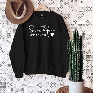 May include: Black sweatshirt with white text that reads "Sweater Weather" with a small white heart. The sweatshirt is hanging on a wooden hanger against a white background with a black and white geometric pattern.