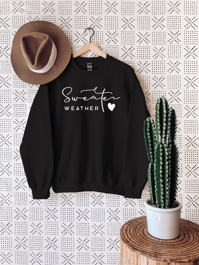 May include: Black sweatshirt with white text that reads "Sweater Weather" with a small white heart. The sweatshirt is hanging on a wooden hanger against a white background with a black and white geometric pattern.