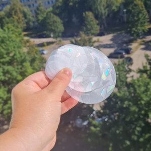 May include: A stack of clear, round, decorative window clings with a geometric design. The clings have a subtle iridescent effect, reflecting a rainbow of colors. The background shows a green, leafy environment.