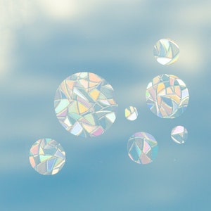 May include: Iridescent, stained-glass-style circular decals in various sizes. The decals are composed of geometric shapes in pastel colors, creating a rainbow effect. They are set against a light blue background, resembling a sky.