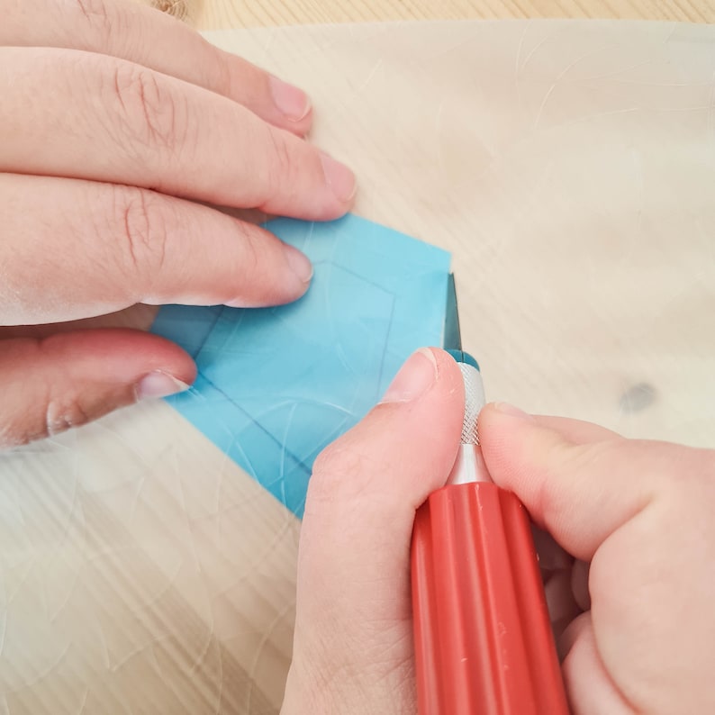 May include: A person's hands using a red craft knife to cut a blue, geometric-shaped sticker on a light-colored surface. The sticker has a faint outline of the shape being cut.