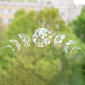 Könnte beinhalten: Ein Fensteraufkleber mit einem Mondphasen-Design. Das Design umfasst einen Vollmond und Halbmonde in verschiedenen Größen. Der Aufkleber hat eine Buntglasoptik mit Pastellfarben und wird vor einem verschwommenen grünen Hintergrund dargestellt.