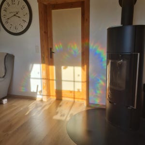 May include: A black modern fireplace with a glass door stands next to a wooden door and a wall clock. The sunlight casts a rainbow pattern on the wall. The room has hardwood floors and a neutral color palette.