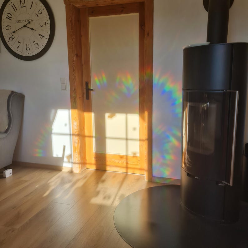 May include: A black modern fireplace with a glass door stands next to a wooden door and a wall clock. The sunlight casts a rainbow pattern on the wall. The room has hardwood floors and a neutral color palette.
