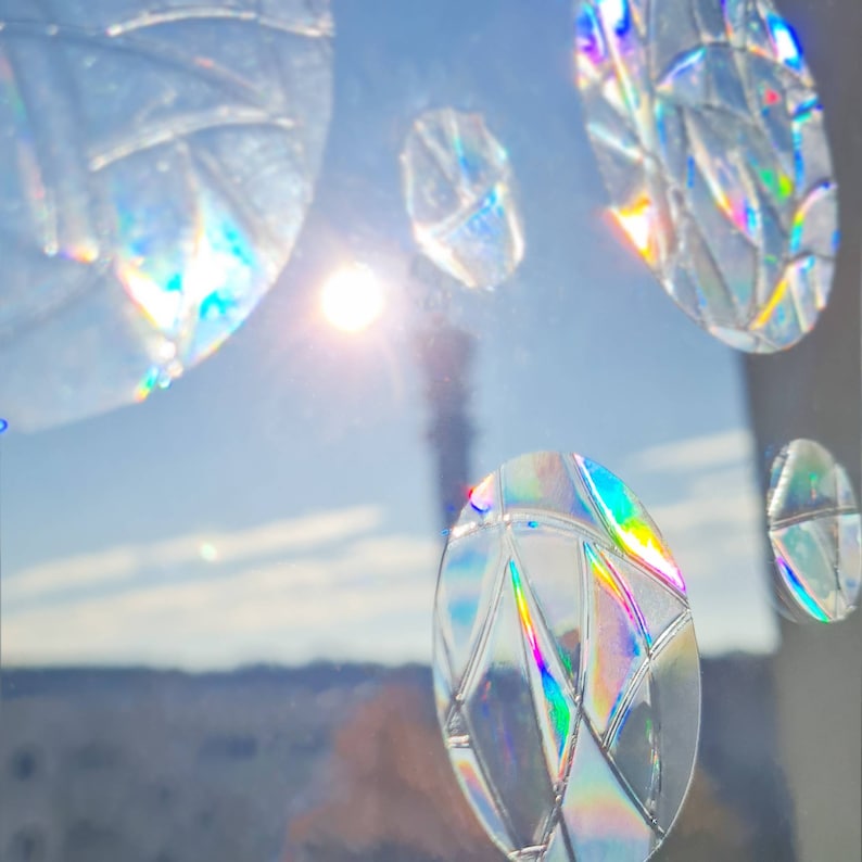 May include: Clear, oval-shaped window clings with a geometric design. The clings have a rainbow iridescent effect, reflecting light with a prism-like quality. The background shows a bright sun and a clear blue sky.