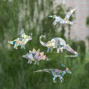 Könnte beinhalten: Fünf bunte Dinosaurier-Fensteraufkleber in verschiedenen Formen, darunter ein Triceratops, ein Stegosaurus und ein Pterodactylus. Die Aufkleber haben einen Glasmalerei-Effekt mit einem Mosaik aus Farben, darunter Blau, Grün, Gelb und Lila.