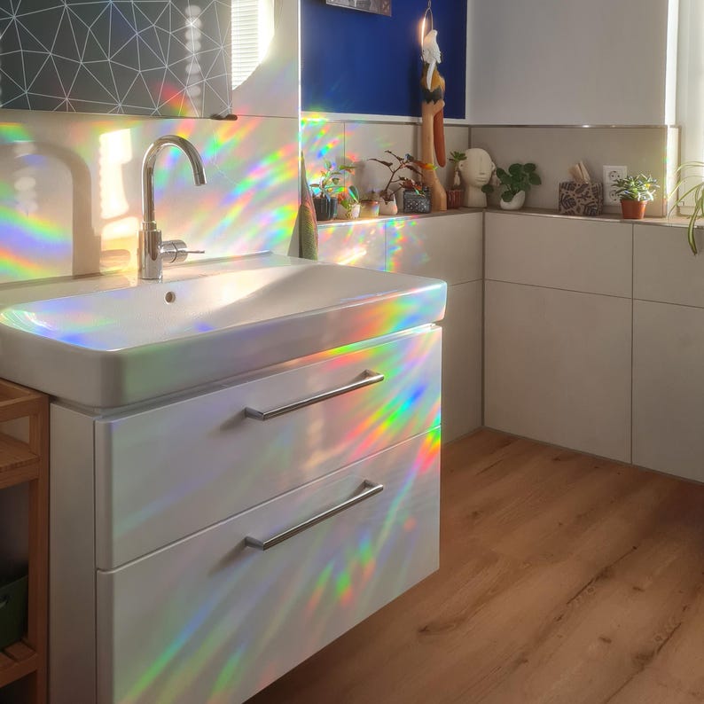 May include: A modern bathroom sink with a chrome faucet and a white cabinet with two drawers. The room is illuminated by rainbow light patterns. Small potted plants and decorative items are on a shelf above the sink.