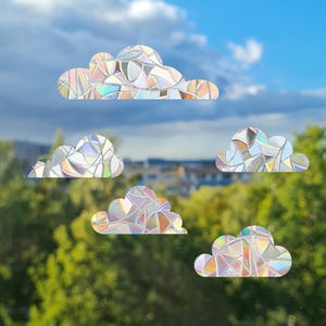 Könnte beinhalten: Fünf wolkenförmige Fensteraufkleber mit Mosaikdesign. Die Aufkleber haben einen regenbogenfarbenen, schillernden Effekt, der verschiedene Farben reflektiert. Die Wolken sind vor dem Hintergrund eines blauen Himmels und grünen Laubes positioniert.