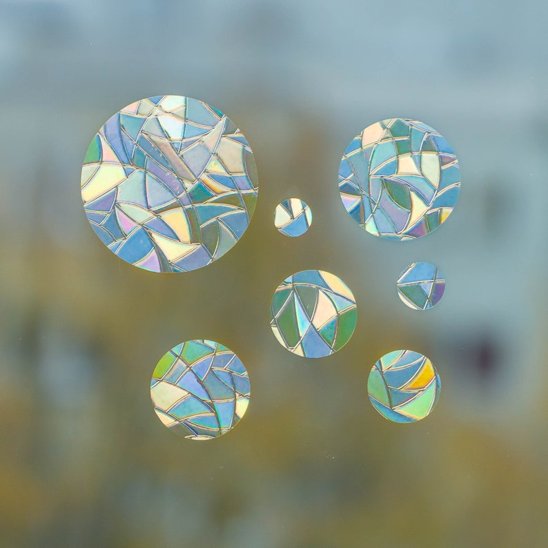 May include: A collection of iridescent, circular window clings. Each cling is composed of irregular, geometric shapes in shades of blue, green, and yellow. The clings vary in size and are arranged in a scattered pattern.