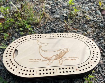 Könnte beinhalten: Cribbage-Brett aus Holz mit Wal-Gravur. Das ovale Brett hat zahlreiche Löcher für die Stifte und einen Schiebedeckel. Das Wal-Design ist in das hellfarbene Holz geätzt, mit den Wörtern "END" und "START" sichtbar.