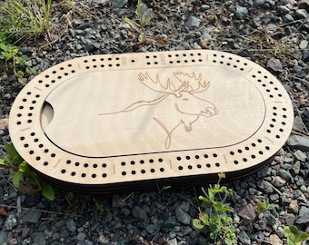 Könnte beinhalten: Ein ovales Cribbage-Brett aus Holz mit einer Elch-Illustration. Das Brett hat eine helle Holzoberfläche mit einem Rand aus Bohrlöchern für die Stifte. Der Elch ist im Stil einer Strichzeichnung dargestellt.