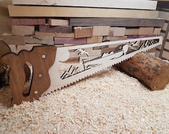 May include: A wooden saw with a detailed cutout design of a mountain scene. The saw is made of light wood and has a brown wooden handle.