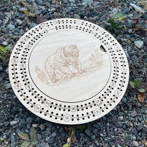 Può includere: Un gioco da tavolo di cribbage rotondo in legno con un disegno di orso grizzly inciso al laser. Il gioco presenta fori numerati per l'inserimento dei pioli e una pista per il punteggio. Il gioco ha un diametro di circa 30 cm.