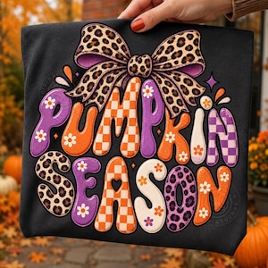 Può includere: Una t-shirt nera con una grande grafica che riporta il testo PUMPKIN SEASON in un carattere arrotondato. Il design include un fiocco con stampa leopardata, motivi a scacchi arancioni e bianchi, accenti floreali viola e arancioni e piccole margherite bianche.