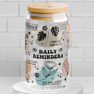May include: A clear glass water bottle with a bamboo lid and a straw. The bottle has a variety of colorful abstract shapes and floral designs with the text "Daily Reminders" and inspirational quotes like "Courage to set boundaries, don't be sorry" and "You have always been enough".