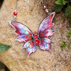 May include: A vibrant butterfly pendant necklace with a silver chain. The butterfly features red, pink, and blue wings, a silver outline, and red gemstone embellishments. The necklace is shown on a rock.