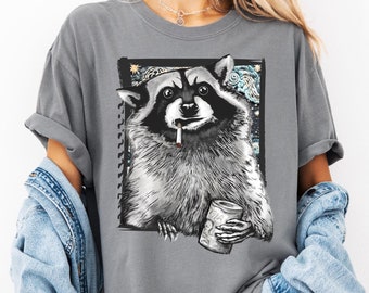 May include: A gray t-shirt with a black and white illustration of a raccoon holding a can and smoking a cigarette. The raccoon is in front of a starry night sky background.