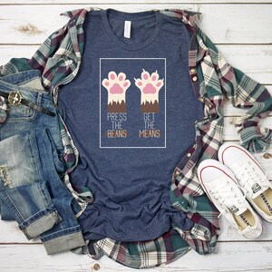 May include: A navy blue t-shirt featuring a graphic of two pink cat paws. The text "Press the Beans" and "Get the Means" is printed below the paws.