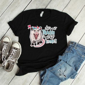 May include: Black t-shirt featuring a cartoon opossum wearing a floral crown, with the text "Let's eat trash and get hit by a car." The shirt is paired with gray sneakers and blue jeans, creating a casual, everyday look.