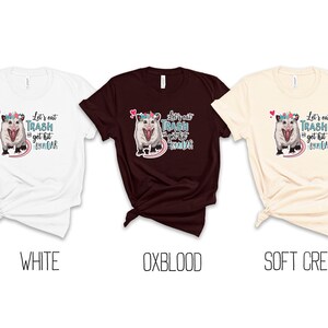 May include: Three t-shirts in white, oxblood, and soft cream. Each shirt features a graphic of an opossum wearing a floral crown, with the text "Let's eat trash & get hit by a car."