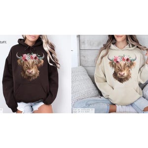 Puede incluir: Dos mujeres con sudaderas con capucha con un dise&ntilde;o de vaca Highland coronada con flores. La sudadera con capucha de la izquierda es de color marr&oacute;n chocolate oscuro y la sudadera con capucha de la derecha es de color arena.