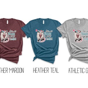 May include: Three t-shirts in heather maroon, heather teal, and athletic grey. Each shirt features a graphic of an opossum wearing a floral crown, with the text "Let's eat trash and get hit by a car."