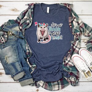 May include: A navy blue t-shirt with a graphic of an opossum wearing a flower crown and the text "Let's eat trash and get hit by a car." The shirt is styled with a plaid shirt, jeans, a belt, and white sneakers.