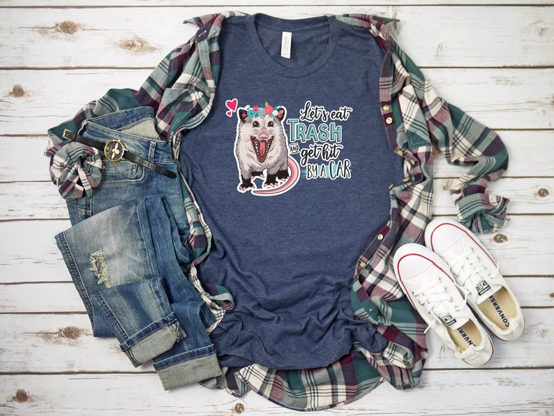 May include: A navy blue t-shirt with a graphic of an opossum wearing a flower crown and the text "Let's eat trash and get hit by a car." The shirt is styled with a plaid shirt, jeans, a belt, and white sneakers.