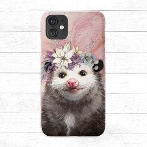 May include: A pink and white phone case with a cute illustration of an opossum wearing a flower crown. The opossum has a pink nose and is looking directly at the camera.