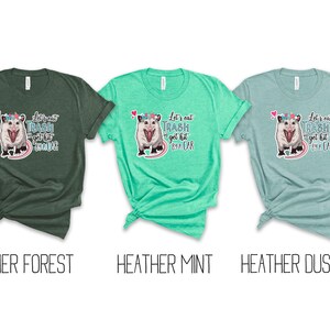 May include: Three heathered t-shirts in forest green, mint, and dusty blue. Each shirt features a graphic of an opossum with a floral crown and the text "Let's eat trash get hit by a car".