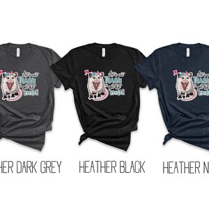 May include: Three t-shirts in heather dark grey, heather black, and heather navy. Each shirt features a graphic of an opossum with a unicorn horn and the text "let's eat trash and get hit by a car".