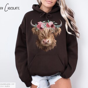 Puede incluir: Una sudadera con capucha de color marr&oacute;n chocolate oscuro con un gr&aacute;fico de vaca de las tierras altas con una corona de flores. El texto "Dark Chocolate" est&aacute; impreso en la parte superior de la sudadera con capucha.