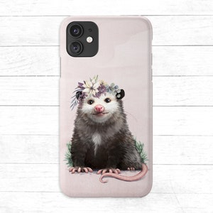 May include: A pink phone case with a cute illustration of a possum wearing a flower crown. The possum is sitting on a bed of green leaves and has a pink nose.
