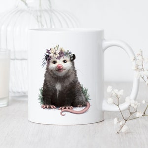 May include: White ceramic mug with a floral crown on a cute opossum. The opossum is sitting on a green leafy background.
