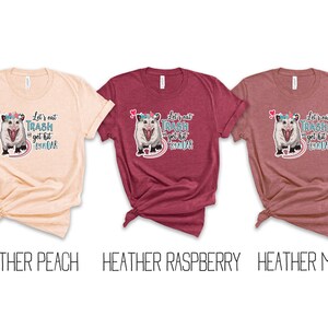 May include: Three t-shirts in heather peach, raspberry, and mauve. Each shirt features a graphic of an opossum with a unicorn horn and the text "Let's eat trash and get hit by a car."