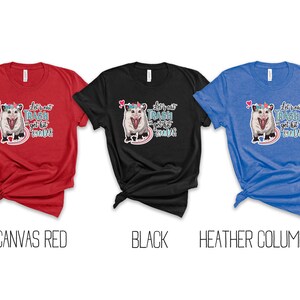 May include: Three t-shirts in red, black, and blue. Each shirt features a graphic of an opossum with a unicorn horn and the text "Let's eat trash and get hit by a car".