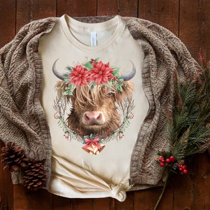 K&ouml;nnte beinhalten: Cremefarbenes T-Shirt mit einer Illustration einer Highland-Kuh. Die Kuh tr&auml;gt einen Blumenkranz mit roten Weihnachtssternen und Gr&uuml;nzeug. Ein brauner Zopfstrick-Cardigan und Tannenzapfen erzeugen ein festliches, gem&uuml;tliches Thema.
