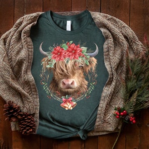 K&ouml;nnte beinhalten: Dunkelgr&uuml;nes T-Shirt mit einem Highland-Kuh-Design, verziert mit einem Kranz aus roten Weihnachtssternen und Gr&uuml;nzeug. Die Kuh hat langes, braunes Fell und gebogene H&ouml;rner. Ein Kiefernzweig und eine braune Strickjacke liegen auf einer Holzoberfl&auml;che.