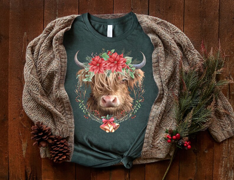 K&ouml;nnte beinhalten: Dunkelgr&uuml;nes T-Shirt mit einem Highland-Kuh-Design, verziert mit einem Kranz aus roten Weihnachtssternen und Gr&uuml;nzeug. Die Kuh hat langes, braunes Fell und gebogene H&ouml;rner. Ein Kiefernzweig und eine braune Strickjacke liegen auf einer Holzoberfl&auml;che.