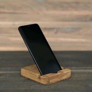 May include: A black smartphone is propped up in a wooden phone stand. The stand is made of light brown wood and has a simple design.