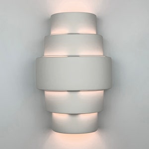 May include: White ceramic wall sconce with five tiers, each tier is a slightly larger circle than the one above it. The sconce is lit from within, casting a warm glow.