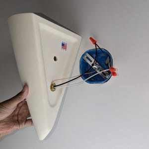 May include: A white ceramic light fixture with a triangular shape and a brass electrical outlet. The fixture is partially attached to a white wall with a blue electrical box and red wire connectors.