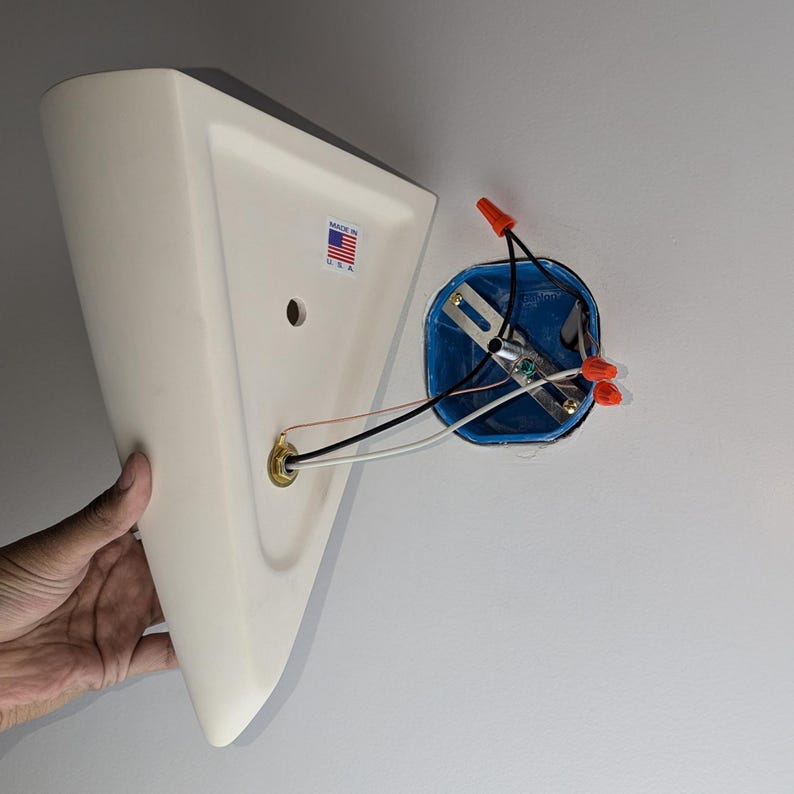 May include: A white ceramic light fixture with a triangular shape and a brass electrical outlet. The fixture is partially attached to a white wall with a blue electrical box and red wire connectors.
