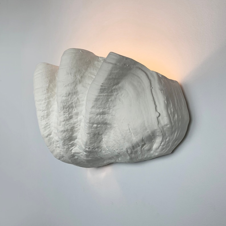 May include: White wall sconce with a textured, organic shape resembling a seashell. The sconce is made of plaster and has a modern design.