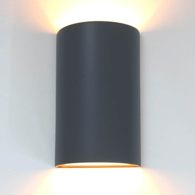 K&ouml;nnte beinhalten: Eine graue zylindrische Wandleuchte mit einem warmwei&szlig;en Licht, das von unten ausstrahlt.