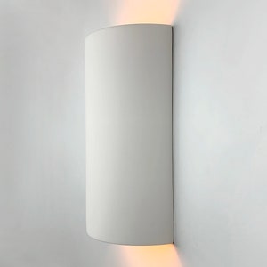 K&ouml;nnte beinhalten: Wei&szlig;e zylindrische Wandleuchte mit abgerundeter Ober- und Unterseite. Die Leuchte ist an einer wei&szlig;en Wand montiert und strahlt ein warmwei&szlig;es Licht aus.