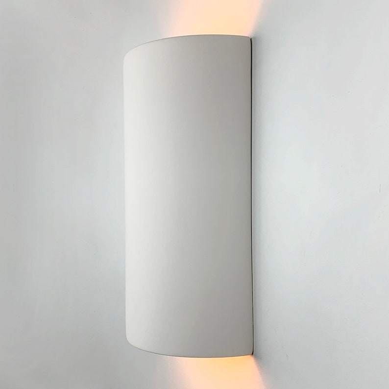 K&ouml;nnte beinhalten: Wei&szlig;e zylindrische Wandleuchte mit abgerundeter Ober- und Unterseite. Die Leuchte ist an einer wei&szlig;en Wand montiert und strahlt ein warmwei&szlig;es Licht aus.