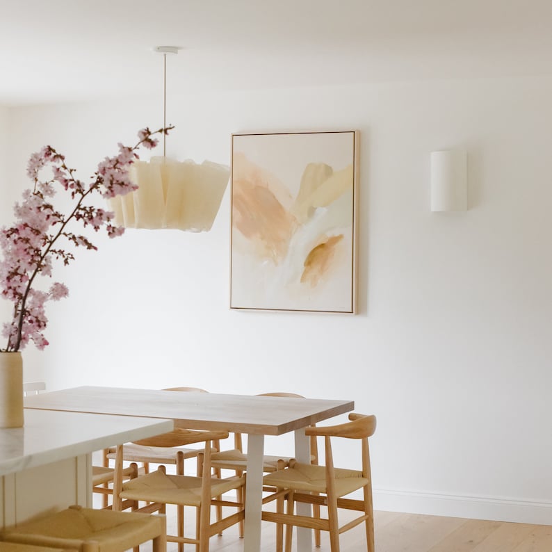 K&ouml;nnte beinhalten: Ein heller Holz-Esstisch mit sechs St&uuml;hlen, einer wei&szlig;en Pendelleuchte und einem hellbraunen abstrakten Gem&auml;lde an der Wand. Eine Vase mit rosa Blumen steht auf dem Tisch.