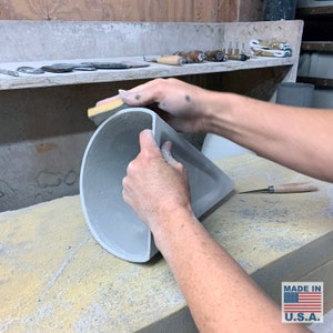 May include: A person's hands are sanding the edge of a gray ceramic planter with a yellow sanding block. The planter is sitting on a light brown surface. The text "MADE IN U.S.A." is visible in the lower right corner of the image.