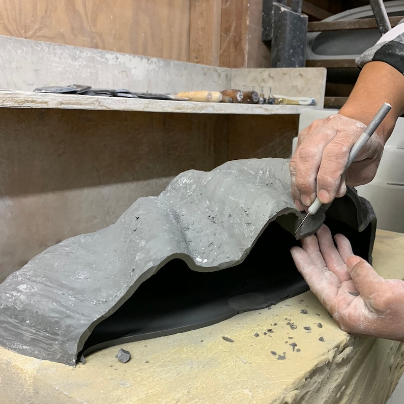 May include: A person is using a small tool to carve a gray ceramic sculpture. The sculpture is shaped like a cave or a mountain range. The sculpture is sitting on a light brown surface.